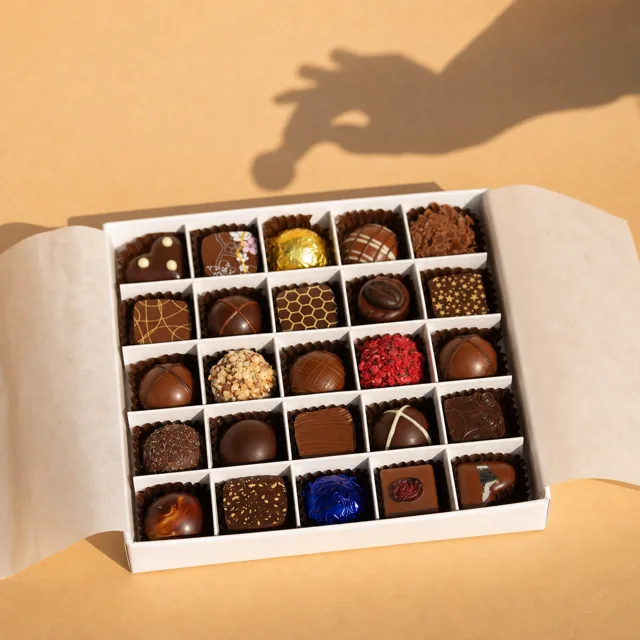 La même boîte de chocolats ouverte, sur un fond ocre, avec l'ombre d'une main