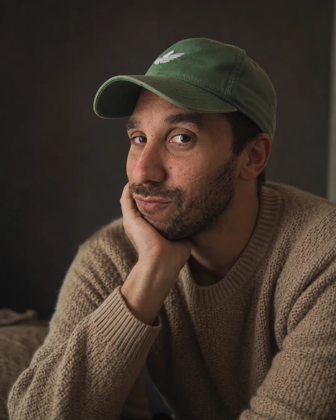 Portrait d'Ismael sur fond sombre, pull en maille beige, casquette verte, la joue posée sur la main