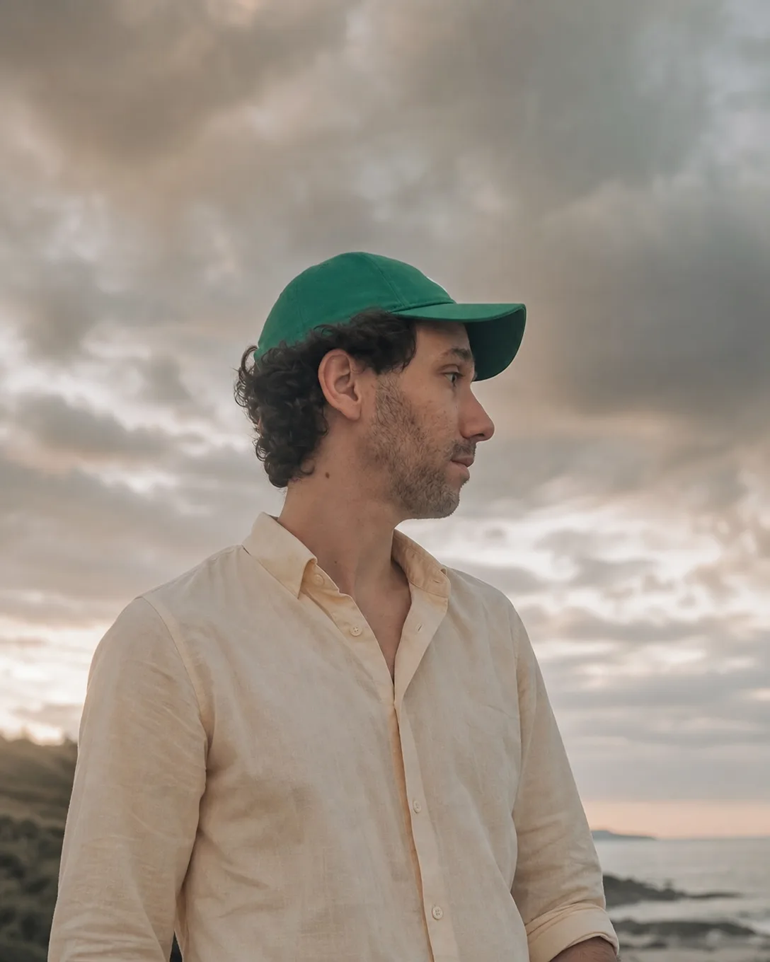 Ismael de profil devant un ciel nuageux au bord de la mer, chemise de lin claire, casquette verte
