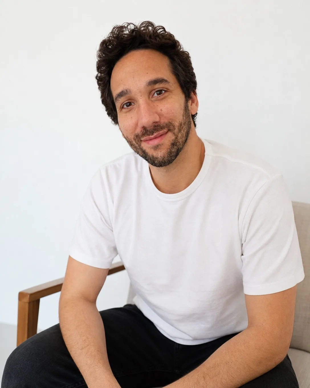 Portrait net d'Ismael assis dans un fauteuil, t-shirt blanc, sur fond clair, sourire