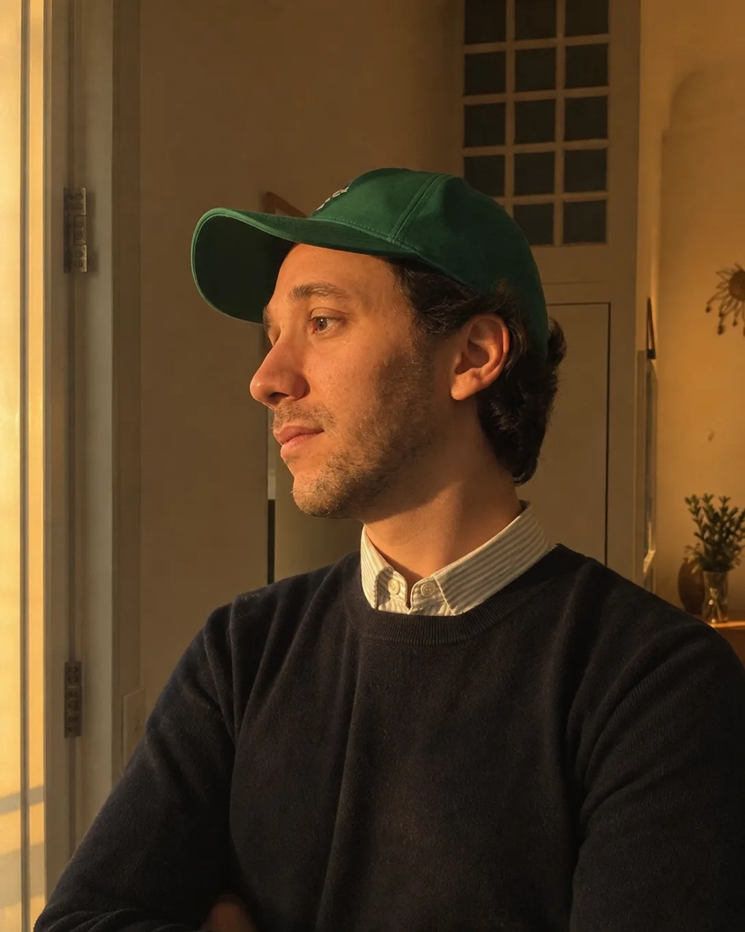 Ismael de profil près d'une fenêtre dans une lumière dorée, pull marine sur une chemise, casquette verte